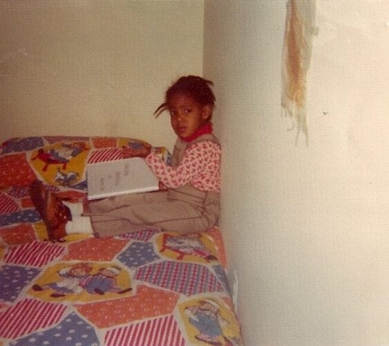 Stacy Ison reading a book, sitting on a bed with Raggedy Ann & Andy sheets. Shoes are on, hair in plaits, annoyed with her mother for disturbing her reading time.