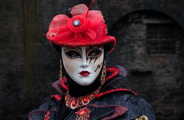 A clown with sad eyes. Face painted white with black around eyes, adorn with a red hat and blue and red coat.