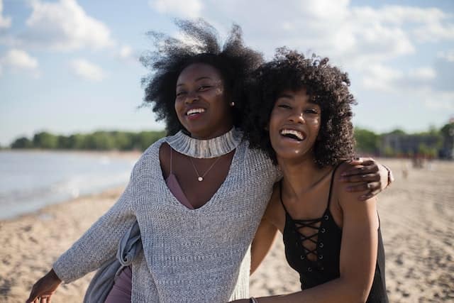 Two women laughing by the ocean. They're both brown-skinned with curly hair. One woman is clad in white, the other in black. They seem like they're best friends in a joyous moment.