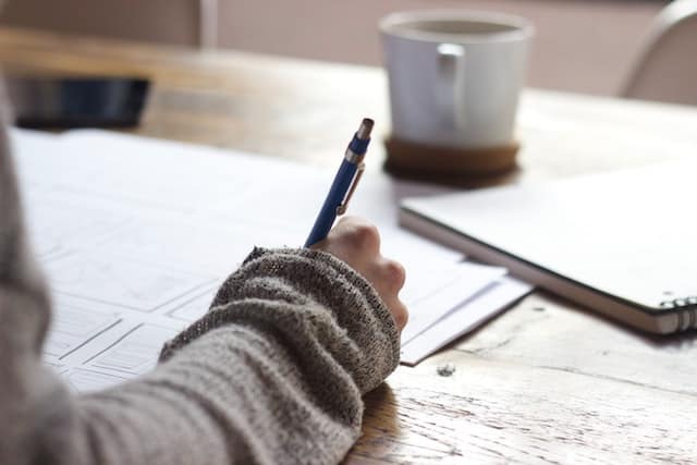 Woman writing with her right hand on a wooden desk. She has a mechanical pencil in her hand and a mug on her desk. She's wearing a long-sleeved gray top. There are different papers on the desk.