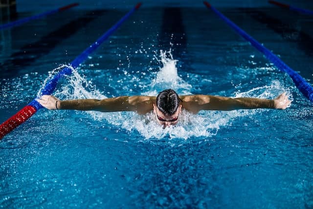 A man is swimming in a blue pool, with goggles on. His arms are stretched out wide, in the middle of a butterfly stroke.