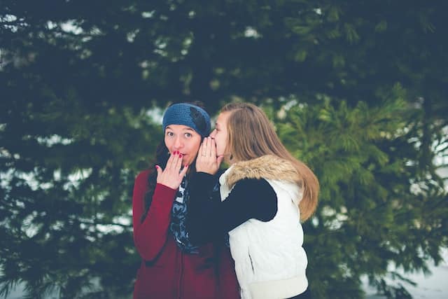 Two women amongst the green trees. It's a cold day. One woman has a blue beanie hat and burgundy sweater. She puts an ungloved hand to her mouth. The other woman, with brown hair, has on a white vest jacket, is whispering in the other woman's ear.