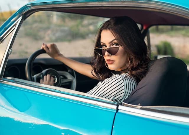 A woman at the wheel of an old-style convertible car. The car is turquoise. She has dark brown, hair. Her hair is down. She has her right hand on 12 o'clock on the wheel. Her sunglasses or on the top of her noise. She's turning to her left, with a mean stare.