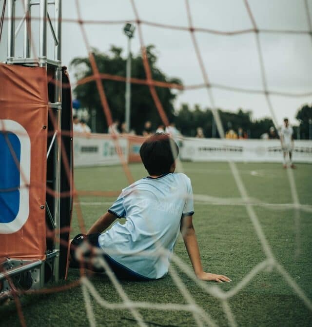 Boy in light blue shirt sitting in front of a soccer net, as if in the goalie, keeper position. The boy has brown hair. There's another player at midfield, standing with a white shirt - maybe ready to take a free kick or start play.