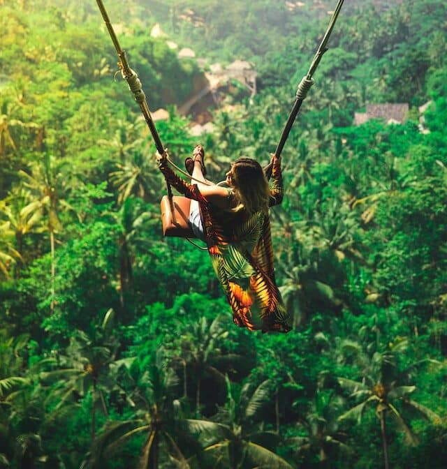 A woman, with brown hair, blonde highlights, sitting on a rope swing in Bali, Indonesia. She's dressed in shorts and a flowy top that is swinging in the air, along with her. She's above many trees, with houses peaking out amongst the foliage.