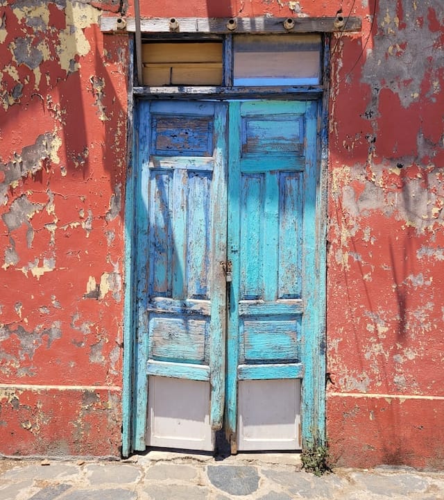 Wabi sabi doors, painted turquoise. Distressed double doors, on a red clay-colored building, where the paint is wearing off.