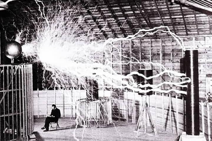Black and white photo of Nikola Tesla sitting in his lab with electrical charges above him. Tesla coil in the lab.