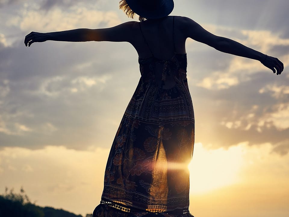 A woman in a halter top, maxi dress, with an arms spread out, free. Short, brown hair is peaking out of a hat. The sun is at dusk. She is on her tippy toes. The light outlines her legs underneath her dress.