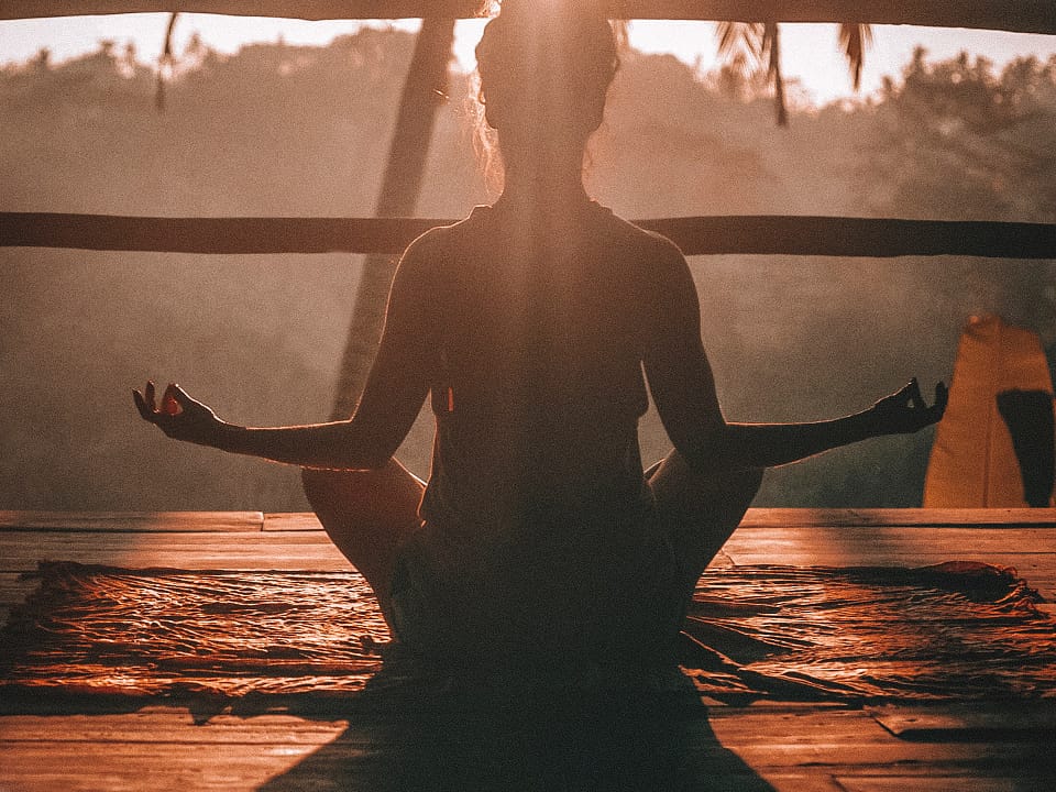 A woman facing the sun, sitting down with her legs crossed, arms are out, palms of hands are open. Yoga or meditation at sunrise with palm trees.