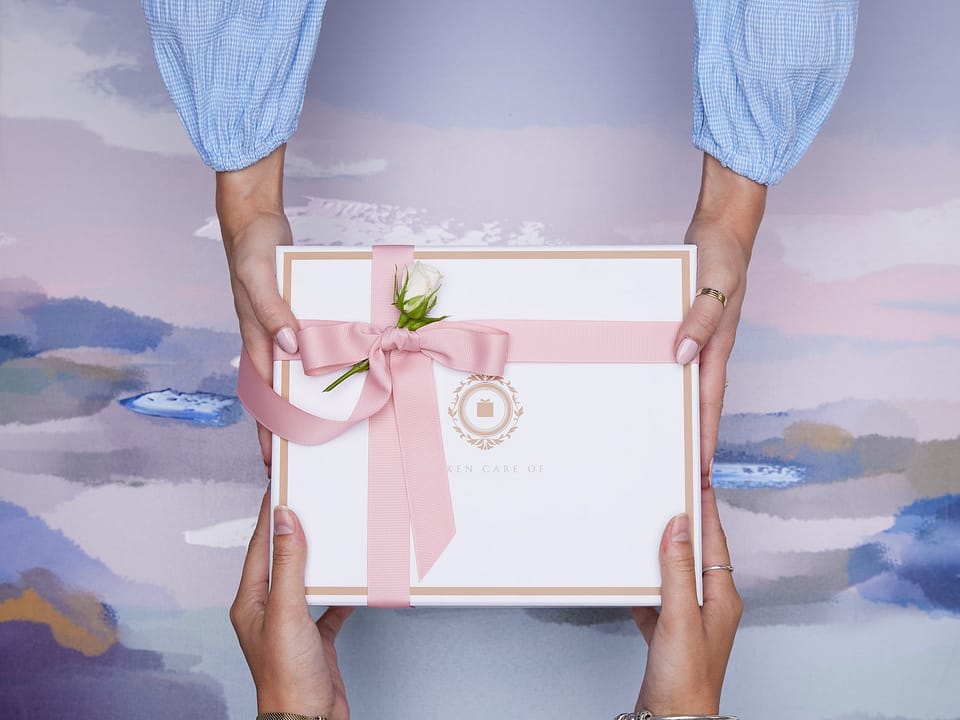 Two people across from each other, each holding a gift in both hands. It's a white box with a pink ribbon. The box has a white flower on it, like a wedding present.