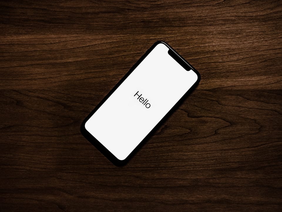 A black iPhone on a wooden desk or table. There's a white screen with the word, "Hello" in black.