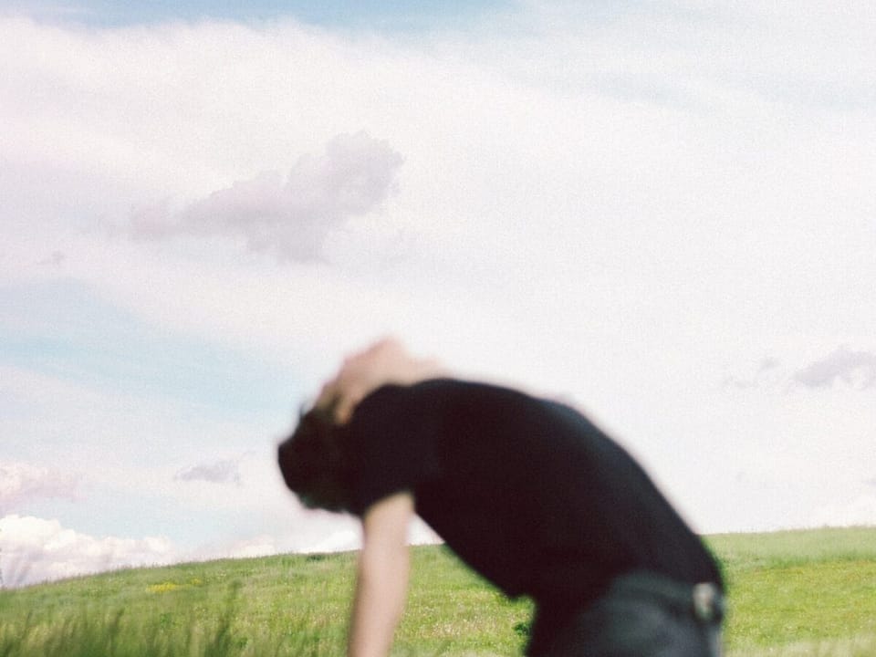 A blurry man in a black shirt and blue jeans, in a filed of grass. He is leaning backwards with his back arched and hands down, away from his body, in an act of surrender.
