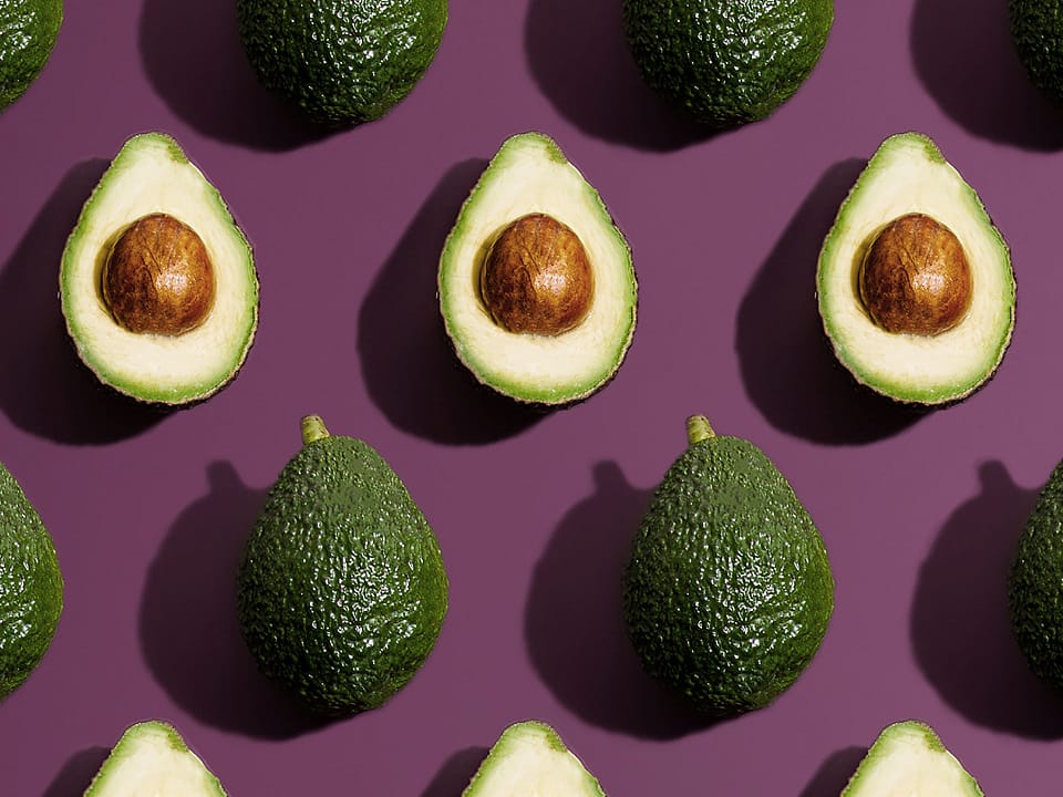 Rows of green avocados, on plum colored background. Some of the avocados are open to look like a woman's womb.
