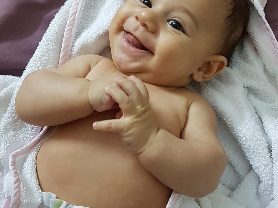 Racially mixed baby in a white, purple, and green diaper, breaking free of a pink and white swaddle, laying on a purple sheet.