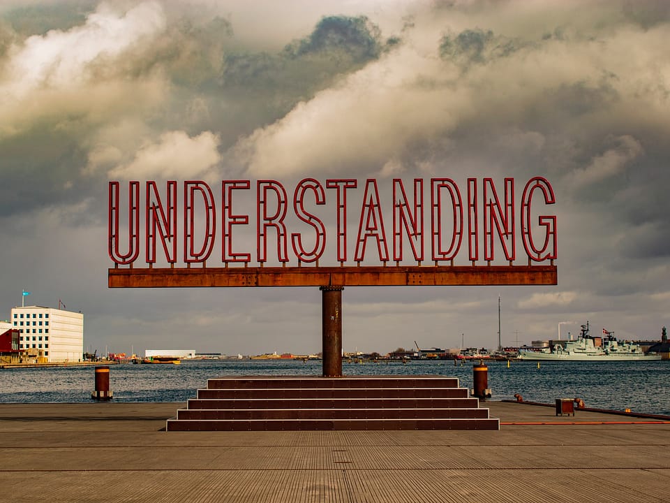 A sign with the word "understanding" in red, on a stained platform, overlooking a body of water, with boats on it.