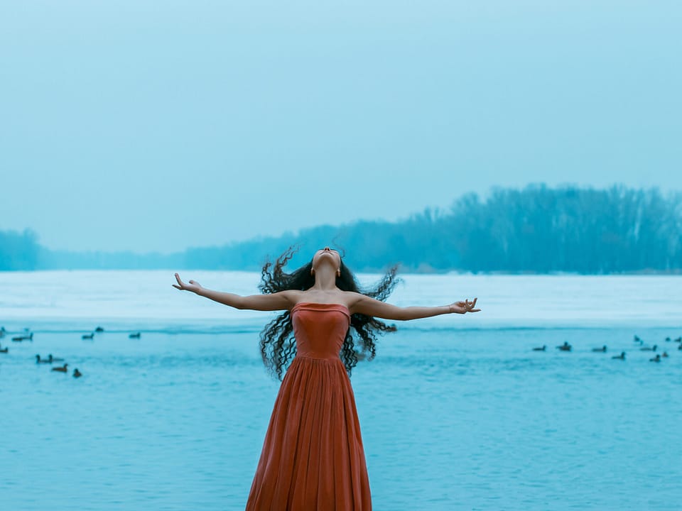 Free goddess woman at the ocean. Beautiful young brunette woman flies with the birds. Background shore of a frozen river. She is wearing a beautiful orange dress. Sad face with a glimmer of hope to escape from the routine of everyday life. Fantasy photo, creative color