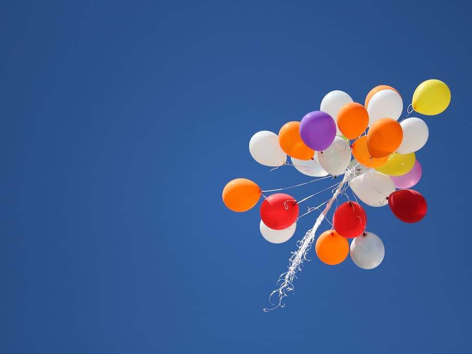A bouquet of balloons, flying in a clear, blue sky. There are white, orange, purple, and red balloons.